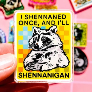 Pode incluir: Um autocolante quadrado amarelo com uma ilustração de guaxinim e o texto "I SHENNANED ONCE, AND I'LL SHENNANIGAN". O guaxinim é preto e branco, com um fundo xadrez azul e amarelo.