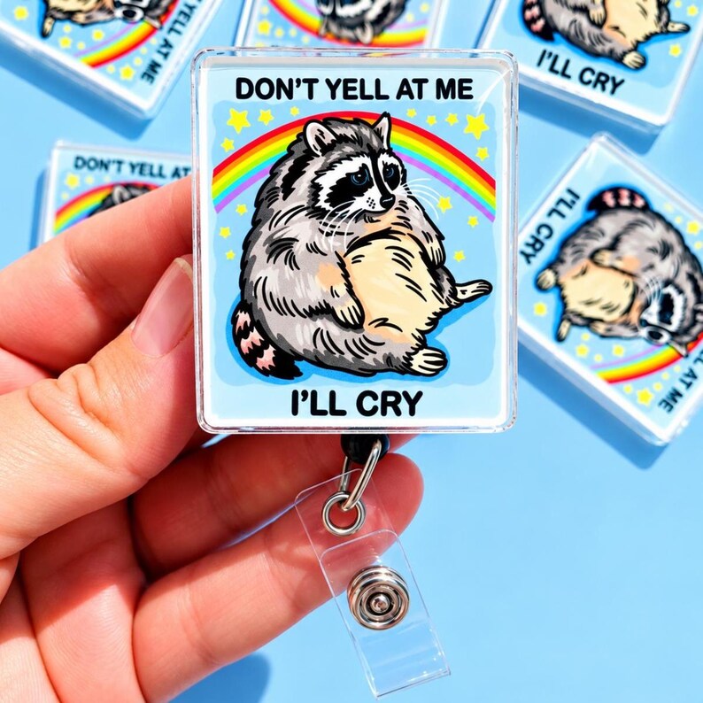 May include: A rectangular badge reel featuring a graphic of a round, grey and black raccoon sitting under a colorful rainbow against a light blue background with yellow stars. The text DON'T YELL AT ME I'LL CRY is printed in black capital letters.