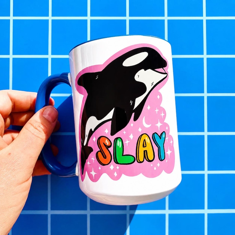 May include: White ceramic mug with a blue handle and rim. The mug features a black and white orca illustration on a pink background with the word "SLAY" in colorful bubble letters. The mug is held by a hand.