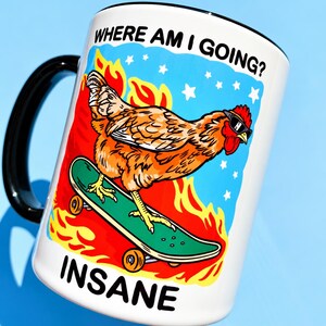 Puede incluir: Una taza de cer&aacute;mica blanca con asa negra que presenta un gr&aacute;fico de una gallina marr&oacute;n con gafas de sol sobre un monopat&iacute;n verde. El fondo muestra llamas naranjas y estrellas blancas sobre un cielo azul. Texto: WHERE AM I GOING? e INSANE.