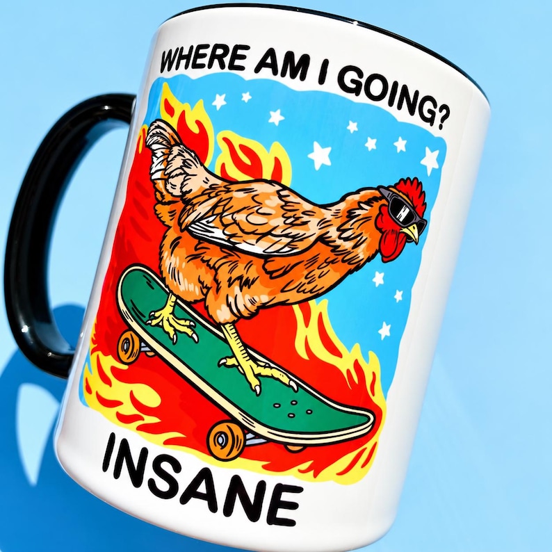 Puede incluir: Una taza de cer&aacute;mica blanca con asa negra que presenta un gr&aacute;fico de una gallina marr&oacute;n con gafas de sol sobre un monopat&iacute;n verde. El fondo muestra llamas naranjas y estrellas blancas sobre un cielo azul. Texto: WHERE AM I GOING? e INSANE.