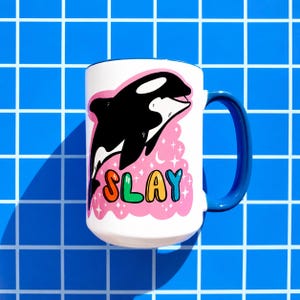 May include: White ceramic mug with a blue handle and rim. The mug features a black and white orca illustration with the word "SLAY" in colorful letters on a pink background with stars.