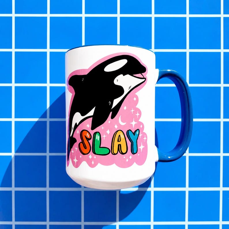 May include: White ceramic mug with a blue handle and rim. The mug features a black and white orca illustration with the word "SLAY" in colorful letters on a pink background with stars.
