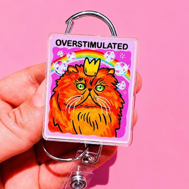 May include: A square acrylic badge reel featuring a graphic of a fluffy orange cat wearing a small yellow crown. Above the cat is a rainbow and the text OVERSTIMULATED in black capital letters. The background is a vibrant pink with bubbles.