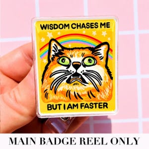 May include: A rectangular badge reel featuring a yellow background with a graphic of an orange cat with wide green eyes. Above the cat is a rainbow and white stars. The text reads WISDOM CHASES ME BUT I AM FASTER in black capital letters.