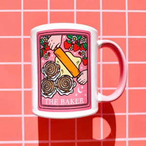 May include: White ceramic mug with a pink handle and a pink border around the design. The design features a baker rolling dough, strawberries, and the text "THE BAKER". The mug is on a pink grid background.