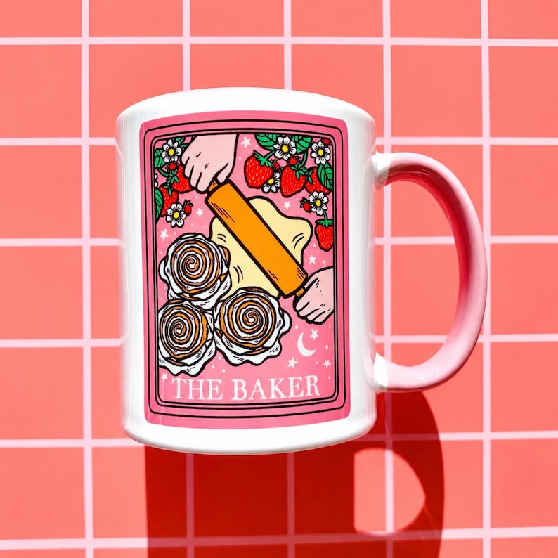 May include: White ceramic mug with a pink handle and a pink border around the design. The design features a baker rolling dough, strawberries, and the text "THE BAKER". The mug is on a pink grid background.