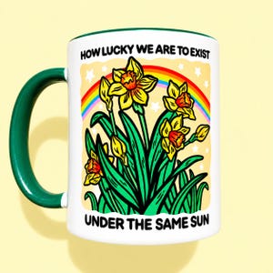 May include: A white ceramic mug with a dark green handle and rim, featuring a colorful illustration of yellow daffodils, green leaves, and a rainbow against a pale yellow background with white stars, and the text HOW LUCKY WE ARE TO EXIST UNDER THE SAME SUN.