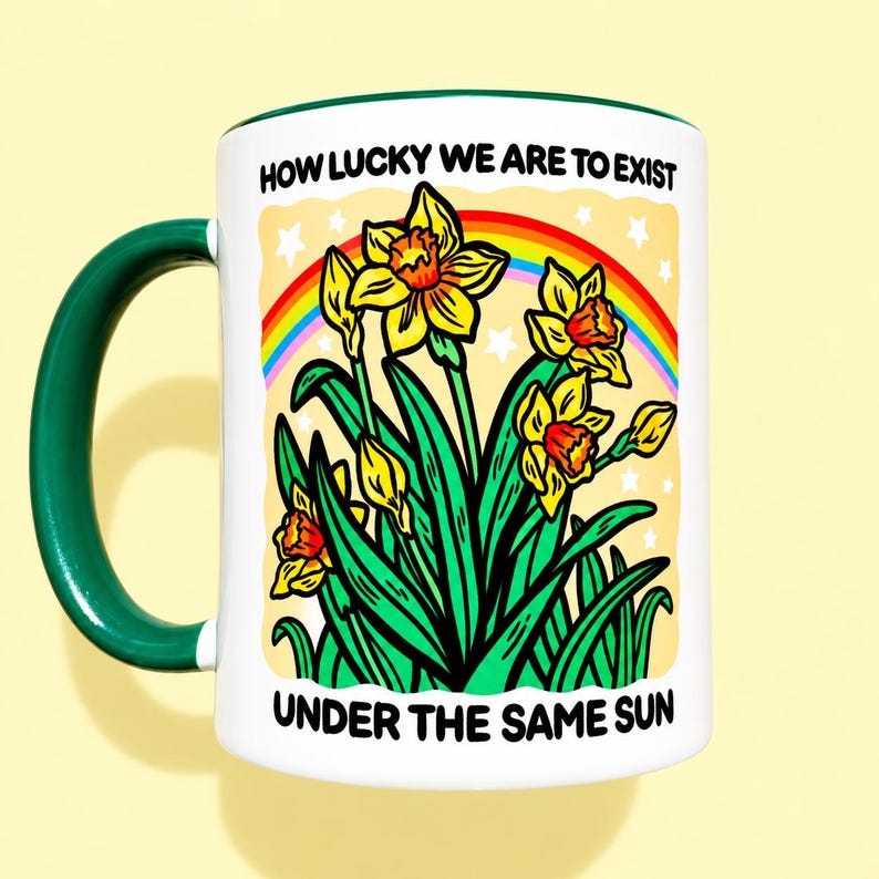 May include: A white ceramic mug with a dark green handle and rim, featuring a colorful illustration of yellow daffodils, green leaves, and a rainbow against a pale yellow background with white stars, and the text HOW LUCKY WE ARE TO EXIST UNDER THE SAME SUN.