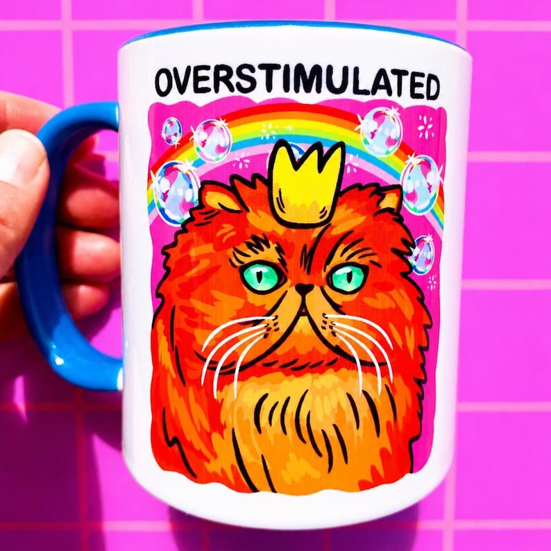 K&ouml;nnte beinhalten: Ein wei&szlig;er Keramikbecher mit blauem Henkel, der eine Grafik einer flauschigen orangefarbenen Katze mit gelber Krone zeigt. &Uuml;ber der Katze befinden sich ein Regenbogen, schwebende Seifenblasen und der Text OVERSTIMULATED in schwarzen Gro&szlig;buchstaben.
