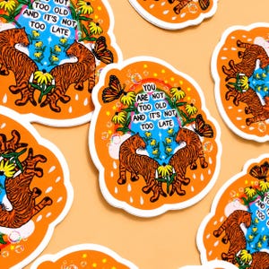 May include: A die-cut sticker featuring two orange tigers facing each other, surrounded by yellow flowers, a butterfly, and a rainbow. The text reads: YOU ARE NOT TOO OLD AND IT'S NOT TOO LATE. The background is a vibrant orange with white raindrop shapes.