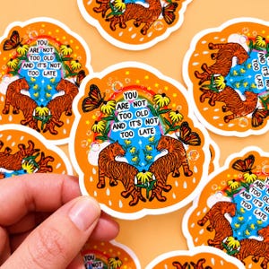 May include: A die-cut sticker featuring two orange tigers facing each other, yellow flowers, a rainbow, butterflies, and bubbles on an orange background. The text reads: YOU ARE NOT TOO OLD AND IT'S NOT TOO LATE. The sticker has a white border.