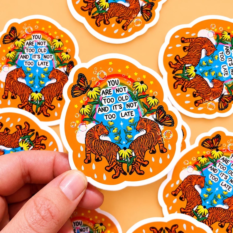 May include: A die-cut sticker featuring two orange tigers facing each other, yellow flowers, a rainbow, butterflies, and bubbles on an orange background. The text reads: YOU ARE NOT TOO OLD AND IT'S NOT TOO LATE. The sticker has a white border.