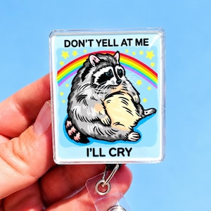 May include: A square badge reel featuring a cartoon illustration of a sad, round raccoon sitting under a rainbow and yellow stars against a light blue background. The text DON'T YELL AT ME and I'LL CRY is printed in black above and below the raccoon.