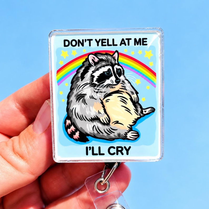 May include: A square badge reel featuring a cartoon illustration of a sad, round raccoon sitting under a rainbow and yellow stars against a light blue background. The text DON'T YELL AT ME and I'LL CRY is printed in black above and below the raccoon.