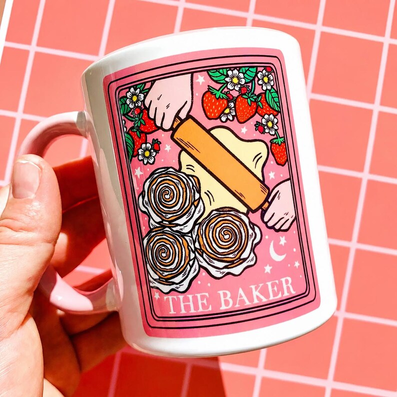 May include: White ceramic mug with a pink handle and a pink rectangular graphic. The graphic shows a rolling pin over dough, strawberries, flowers, and the text "THE BAKER".