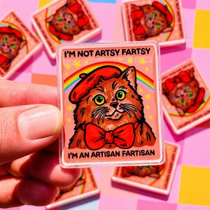 May include: A clear, rectangular sticker featuring a cartoon cat wearing a red beret and bow tie, with the text "I'M NOT ARTSY FARTSY I'M AN ARTISAN FARTISAN" above and below the cat. The cat is brown with green eyes, set against a pink background with a rainbow.