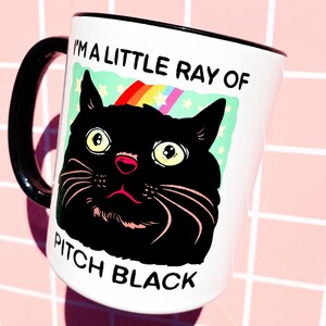 May include: White ceramic mug with a black handle and rim. The mug features a black cat illustration with a rainbow and the text "I'M A LITTLE RAY OF PITCH BLACK". The cat has yellow eyes and a pink nose.