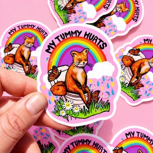 May include: A die-cut sticker featuring a fox sitting on a toilet with the text MY TUMMY HURTS. The design includes a colorful rainbow, monarch butterflies, white daisies, green grass, blue raindrops, and holographic bubbles on a pink background.