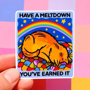 Puede incluir: Una pegatina con un gato naranja de dibujos animados acostado sobre una cama colorida, bajo un arco&iacute;ris y estrellas. El texto "HAVE A MELTDOWN YOU'VE EARNED IT" est&aacute; impreso en la pegatina, sostenida por una mano sobre un fondo rosa y morado.