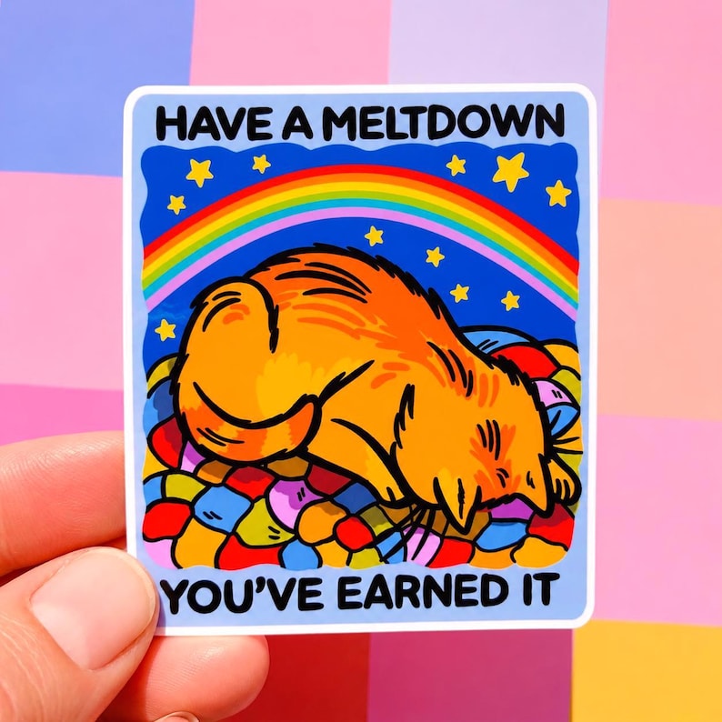 Puede incluir: Una pegatina con un gato naranja de dibujos animados acostado sobre una cama colorida, bajo un arco&iacute;ris y estrellas. El texto "HAVE A MELTDOWN YOU'VE EARNED IT" est&aacute; impreso en la pegatina, sostenida por una mano sobre un fondo rosa y morado.