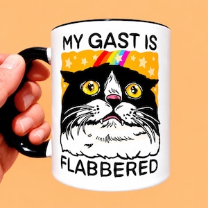 May include: A white ceramic mug with a black handle and rim, featuring a graphic of a black and white cat with wide yellow eyes. Above the cat is the text MY GAST IS and below is FLABBERED, set against an orange background with yellow stars and a rainbow.