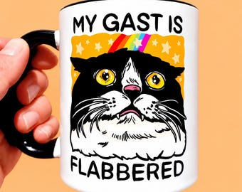May include: A white ceramic mug with a black handle and rim, featuring a graphic of a black and white cat with wide yellow eyes. Above the cat is the text MY GAST IS and below is FLABBERED, set against an orange background with yellow stars and a rainbow.