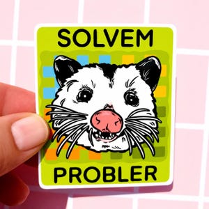 May include: A sticker with a cartoon opossum face and the words "SOLVEM PROBLER" in black capital letters. The opossum has a pink nose, black eyes, and white fur with black accents. The background is green with a grid of blue, orange, and yellow squares.