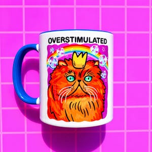 K&ouml;nnte beinhalten: Ein wei&szlig;er Keramikbecher mit blauem Henkel und einer Grafik einer flauschigen orangefarbenen Katze mit gelber Krone. &Uuml;ber der Katze steht der Text OVERSTIMULATED in schwarzen Buchstaben. Ein bunter Regenbogen und Blasen umgeben die Katze.