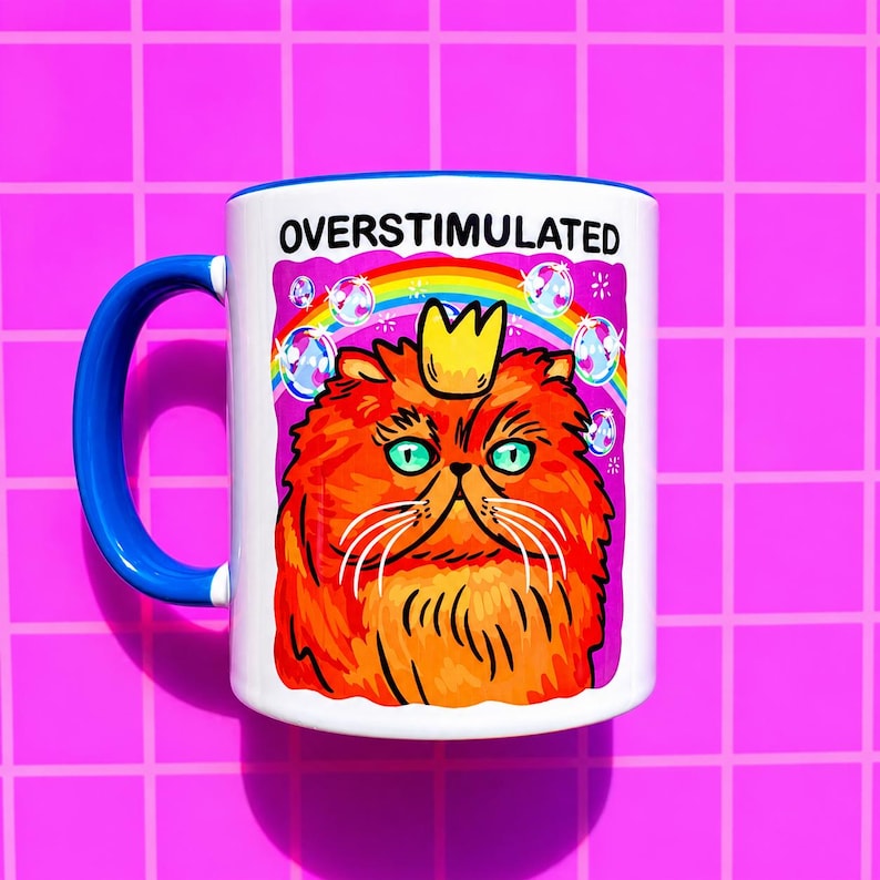 K&ouml;nnte beinhalten: Ein wei&szlig;er Keramikbecher mit blauem Henkel und einer Grafik einer flauschigen orangefarbenen Katze mit gelber Krone. &Uuml;ber der Katze steht der Text OVERSTIMULATED in schwarzen Buchstaben. Ein bunter Regenbogen und Blasen umgeben die Katze.