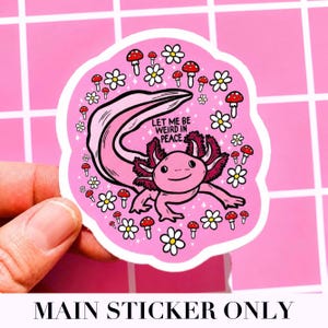 May include: A pink sticker featuring a cartoon axolotl with the text "LET ME BE WEIRD IN PEACE." The axolotl is surrounded by white daisies and red mushrooms. The sticker is held against a pink background.