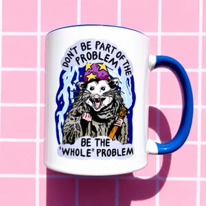 Pu&ograve; includere: Tazza in ceramica bianca con manico e bordo blu. La tazza presenta una grafica di un opossum con un cappello da mago e un bastone, con il testo "DON'T BE PART OF THE PROBLEM BE THE *WHOLE* PROBLEM."