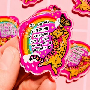 Op de afbeelding: Een doorzichtige acryl pin met een felroze achtergrond waarop een gele kat naar een monarchvlinder reikt. Een kleurrijke regenboog, witte wolken en gele sterren omringen de kat. De tekst luidt: IT'S NOT ALL SUNSHINE & RAINBOWS BUT A GOOD AMOUNT OF IT ACTUALLY IS.