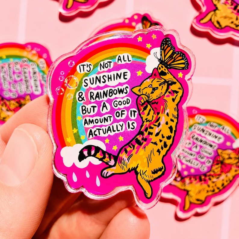 Op de afbeelding: Een doorzichtige acryl pin met een felroze achtergrond waarop een gele kat naar een monarchvlinder reikt. Een kleurrijke regenboog, witte wolken en gele sterren omringen de kat. De tekst luidt: IT'S NOT ALL SUNSHINE & RAINBOWS BUT A GOOD AMOUNT OF IT ACTUALLY IS.