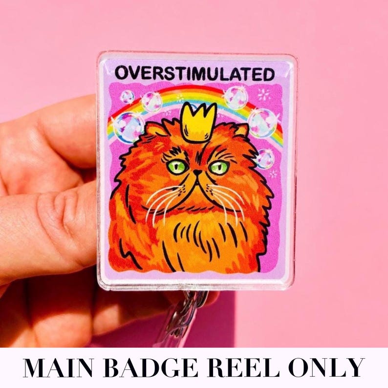 May include: A square badge reel featuring an orange cat with green eyes wearing a yellow crown. The background is light purple with a rainbow and bubbles. The text OVERSTIMULATED is printed in black at the top of the badge reel design.