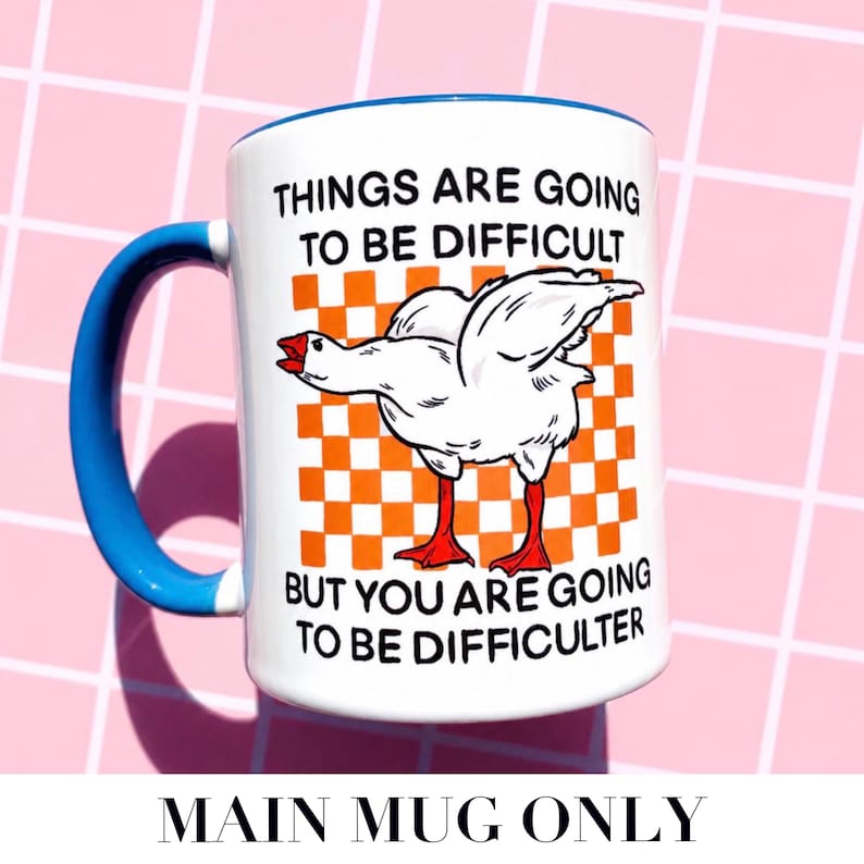May include: White ceramic mug with a blue handle and rim. The mug features a cartoon goose illustration on an orange and white checkered background. Text on the mug reads "THINGS ARE GOING TO BE DIFFICULT BUT YOU ARE GOING TO BE DIFFICULTER."