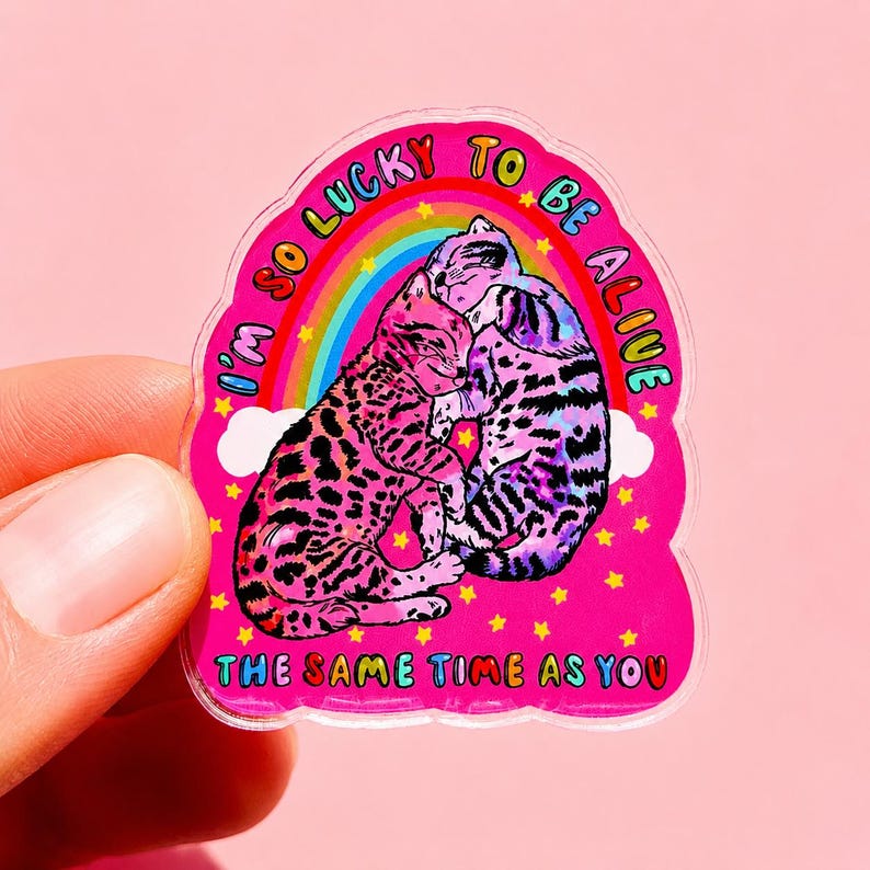 May include: A die-cut sticker featuring two cats cuddling against a rainbow and white clouds on a bright pink background. The text reads I'M SO LUCKY TO BE ALIVE THE SAME TIME AS YOU in colourful letters, surrounded by small yellow stars.