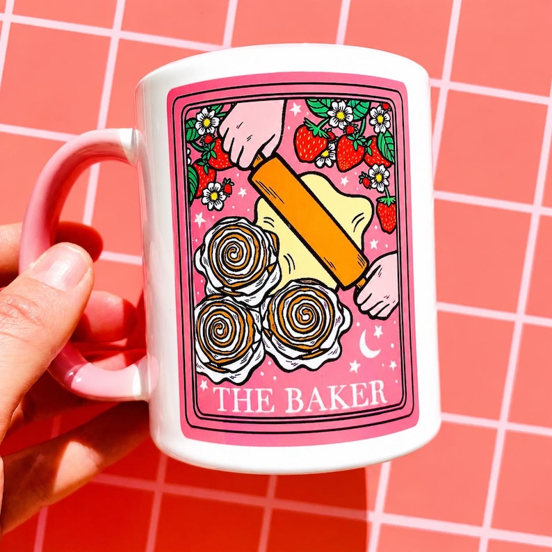 May include: White ceramic mug with a pink handle and border. The mug features a graphic of a baker rolling dough, strawberries, flowers, and the text "THE BAKER".
