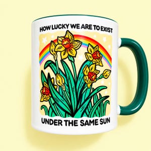 May include: A white ceramic mug with a dark green handle and rim. The graphic features yellow daffodils, a colorful rainbow, and white stars against a pale yellow background. The text reads: HOW LUCKY WE ARE TO EXIST UNDER THE SAME SUN.
