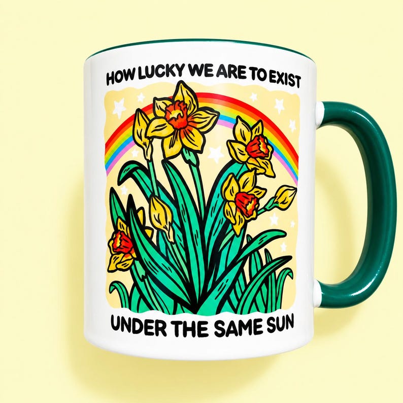 May include: A white ceramic mug with a dark green handle and rim. The graphic features yellow daffodils, a colorful rainbow, and white stars against a pale yellow background. The text reads: HOW LUCKY WE ARE TO EXIST UNDER THE SAME SUN.