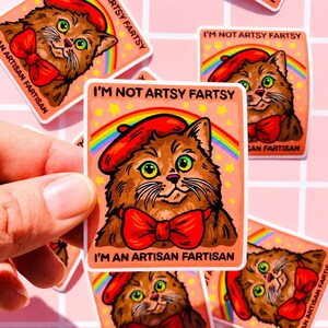 Pu&ograve; includere: Un adesivo quadrato raffigurante un gatto marrone che indossa un basco rosso e un papillon rosso. Dietro il gatto ci sono un arcobaleno e stelle gialle su sfondo color pesca. Il testo recita I'M NOT ARTSY FARTSY e I'M AN ARTISAN FARTISAN.