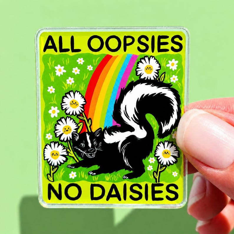 May include: A rectangular sticker featuring a black and white skunk standing in a field of white daisies with yellow centres. A colourful rainbow arches behind the skunk. The text ALL OOPSIES NO DAISIES is printed in black at the top and bottom.