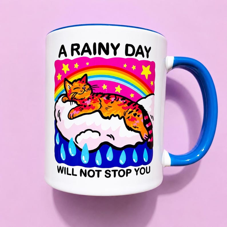 May include: A white ceramic mug with a blue handle and interior. The graphic features an orange cat resting on a white cloud under a rainbow and yellow stars, with blue raindrops below. The text reads A RAINY DAY WILL NOT STOP YOU.
