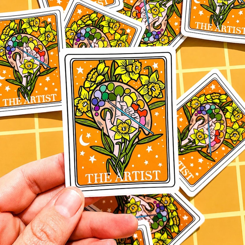 May include: Orange sticker featuring a hand holding a paint palette with a paintbrush, surrounded by yellow daffodils. The text "THE ARTIST" is at the bottom. Stars and a crescent moon decorate the background. The sticker has a white border.