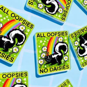 May include: Rectangular acrylic pins featuring a black and white skunk illustration on a green background with white daisies and a rainbow. The text ALL OOPSIES NO DAISIES is printed in black capital letters on each pin, which has a clear finish.