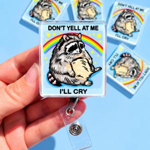 May include: A square badge reel featuring a cartoon raccoon with grey and black fur, a cream-colored belly, and a striped tail. The background is light blue with a colorful rainbow and yellow stars. The text reads DON'T YELL AT ME I'LL CRY.