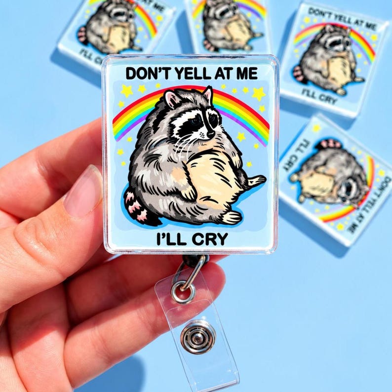 May include: A square badge reel featuring a cartoon raccoon with grey and black fur, a cream-colored belly, and a striped tail. The background is light blue with a colorful rainbow and yellow stars. The text reads DON'T YELL AT ME I'LL CRY.