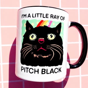 May include: White ceramic mug with a black handle and rim. The mug features a cartoon illustration of a black cat with large yellow eyes, a pink nose, and long white whiskers. Text on the mug reads "I'M A LITTLE RAY OF PITCH BLACK."