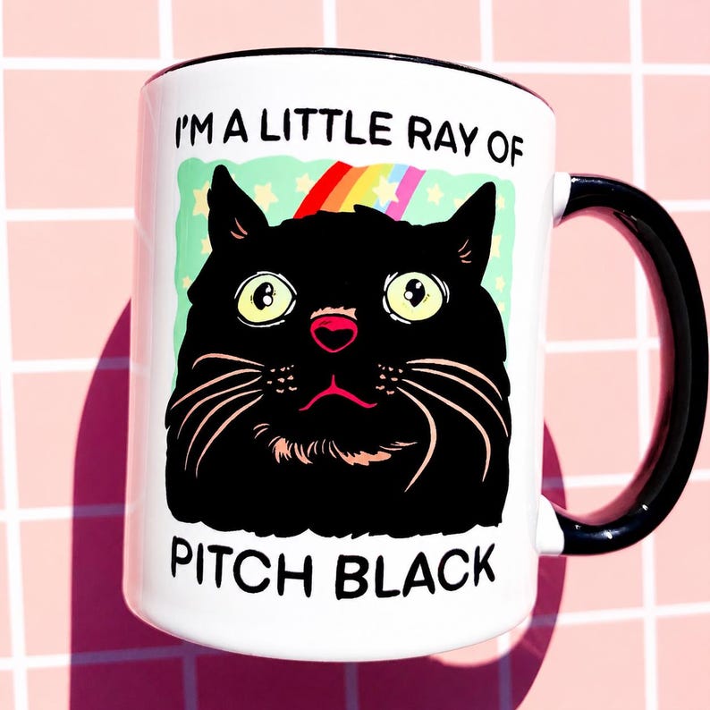 May include: White ceramic mug with a black handle and rim. The mug features a cartoon illustration of a black cat with large yellow eyes, a pink nose, and long white whiskers. Text on the mug reads "I'M A LITTLE RAY OF PITCH BLACK."