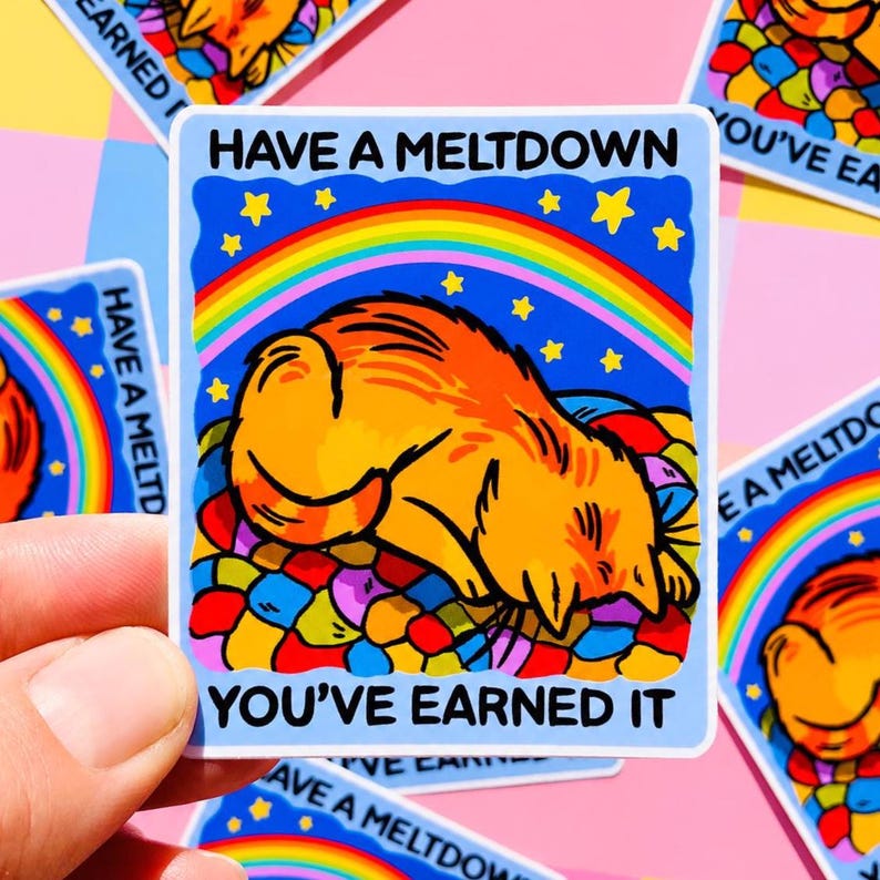 Puede incluir: Una pegatina cuadrada con borde azul presenta un gato naranja de dibujos animados acostado sobre una cama colorida. Encima del gato hay un arco&iacute;ris y estrellas, con el texto "HAVE A MELTDOWN" y "YOU'VE EARNED IT".